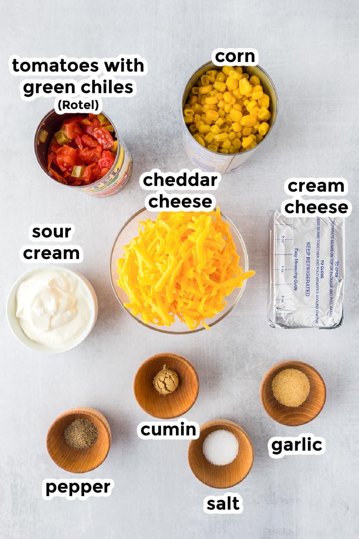 Ingredients arranged on a surface including canned tomatoes with green chiles, corn, shredded cheddar cheese, cream cheese, sour cream, pepper, cumin, salt, and garlic.