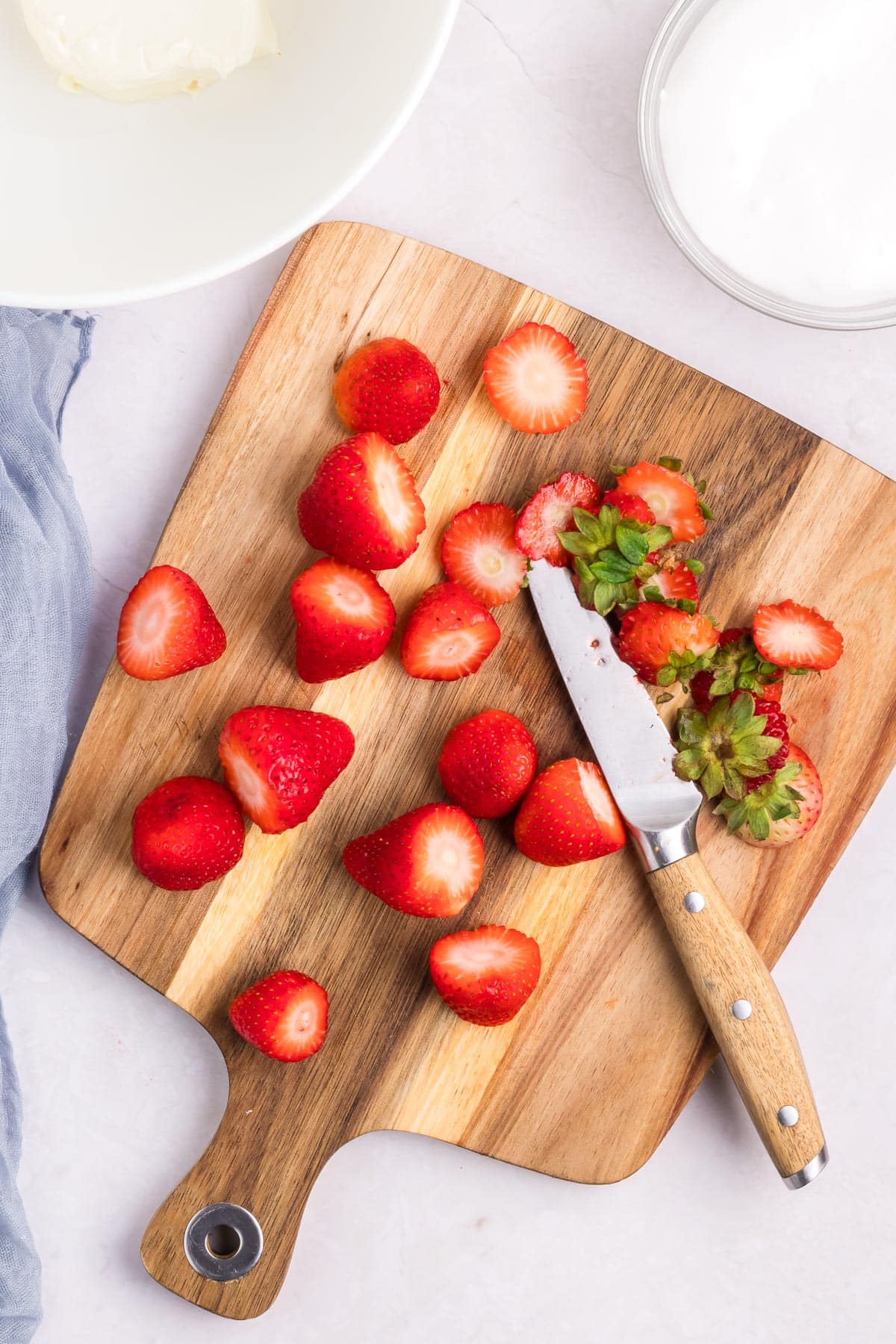 A wooden cutting board holds sliced strawberries and a paring knife, with a bowl and a cloth on the side.