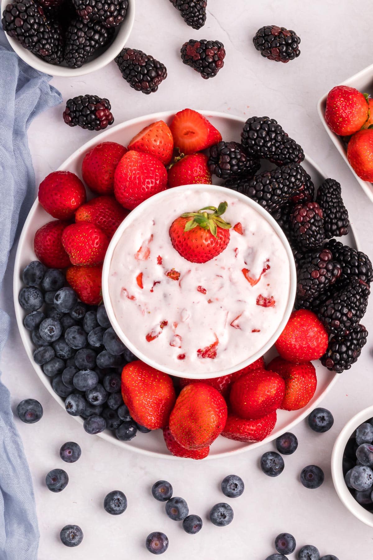 A plate of cream cheese fruit dip with strawberry pieces and a full strawberry in the dip surrounded by a platter of fresh berries.