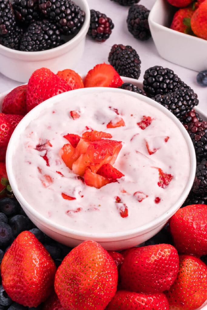 A bowl of creamy fruit dip, garnished with chopped strawberries, surrounded by fresh strawberries, blackberries, and blueberries in and around small dishes.