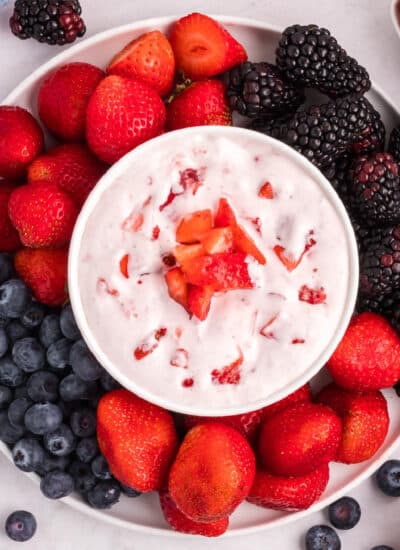 Square view of a cream cheese fruit dip topped with strawberry bits and surrounded by a platter of berries to dip.