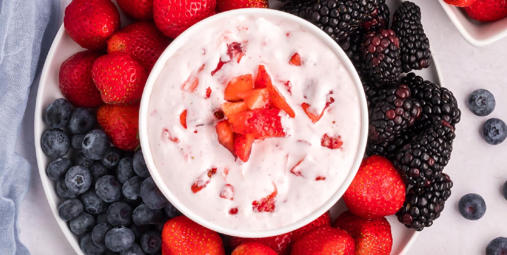 A bowl of cream cheese fruit dip topped with strawberry pieces and fresh fruit on a platter around the dip.