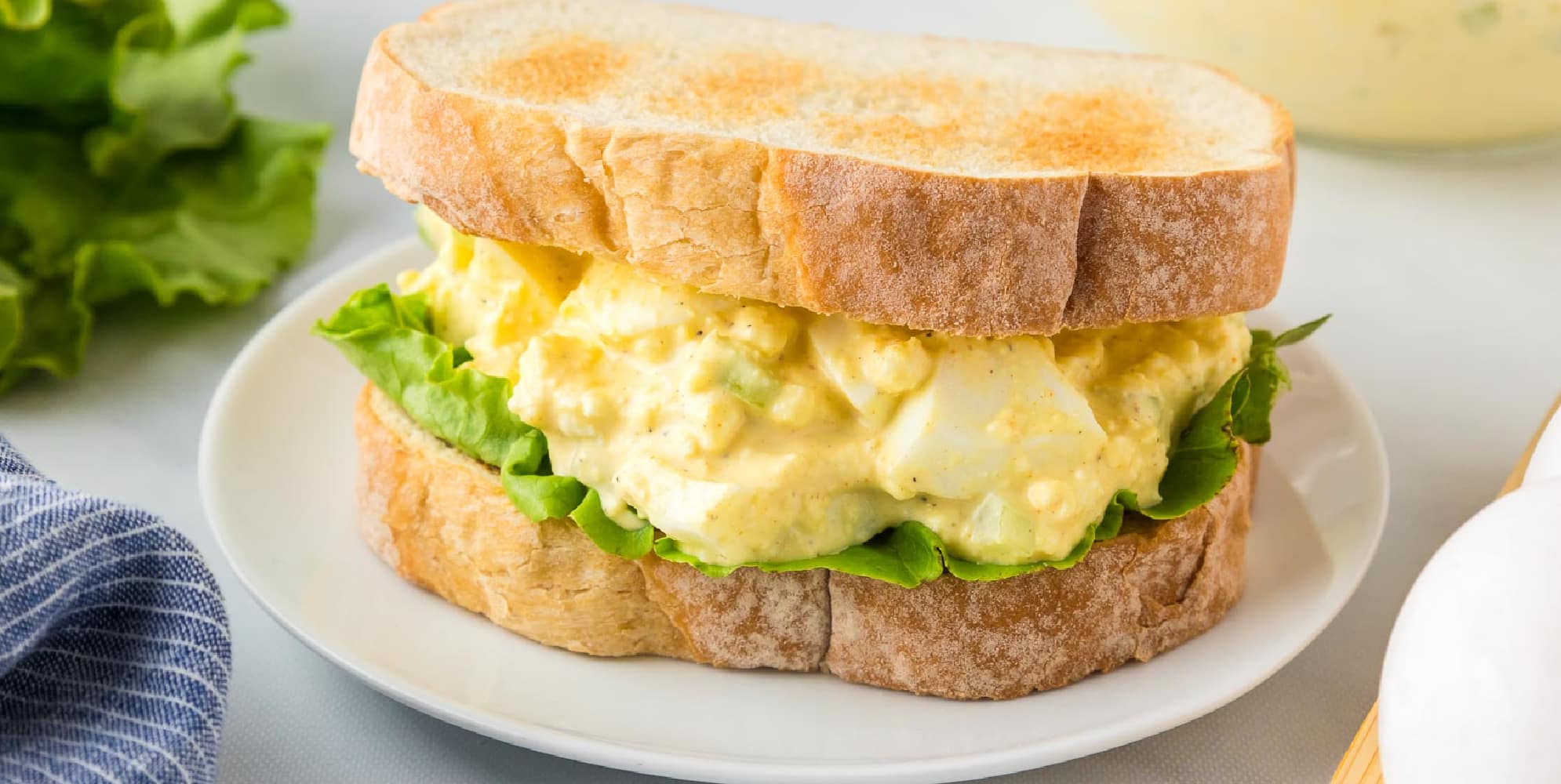 A sandwich on a white plate contains egg salad and lettuce, placed between two slices of toasted bread.