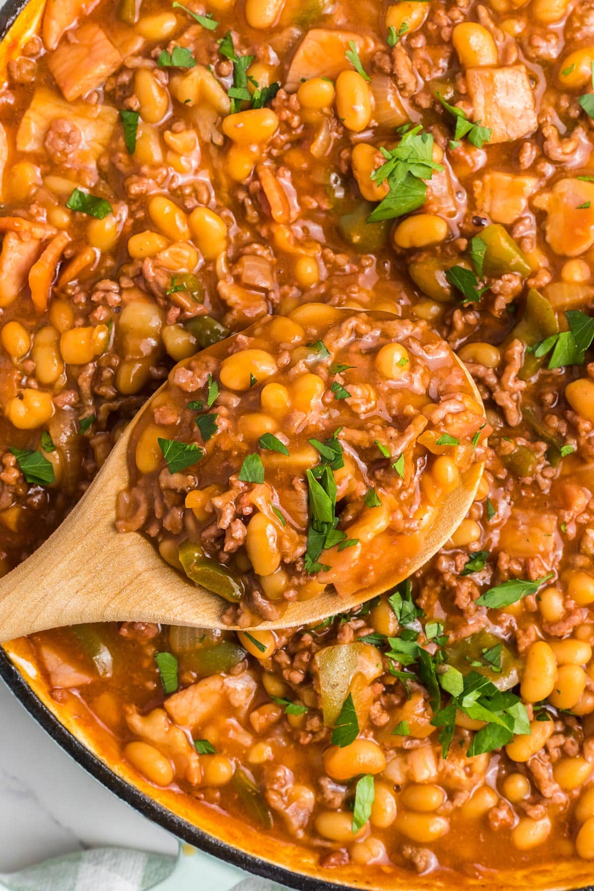 A wooden spoon scoops up a portion of baked beans mixed with ground beef from a large pan.