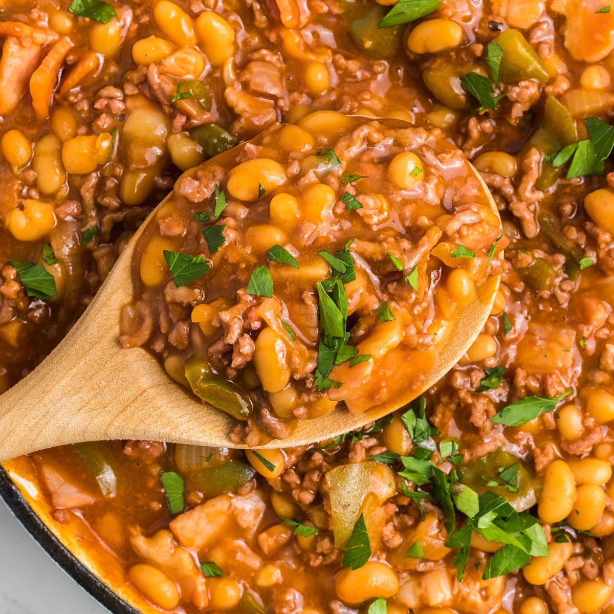 Close-up of a wooden spoon scooping a hearty chili with baked beans, ground meat, and chopped green peppers, garnished with parsley.