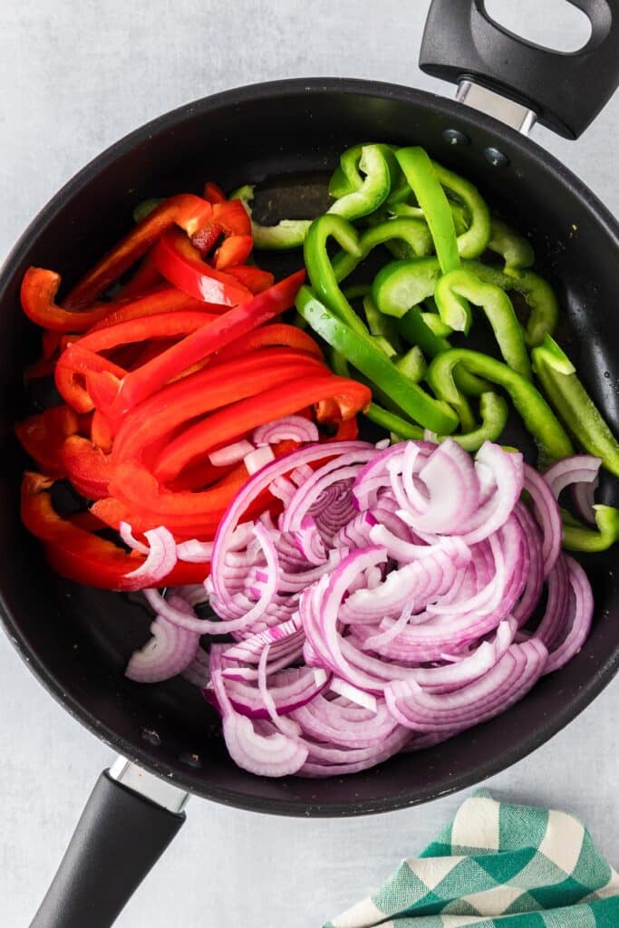 A frying pan with sliced red bell peppers, green bell peppers, and red onions before cooking.