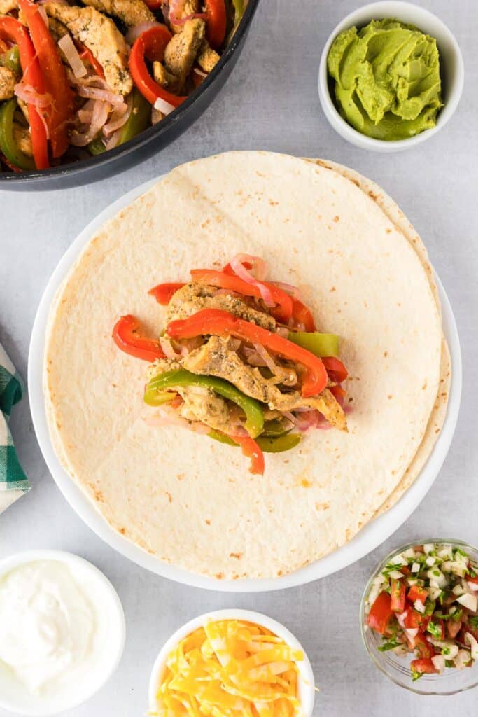 A tortilla filled with cooked chicken, peppers and onions waiting to be filled with other toppings and rolled into a wrap.