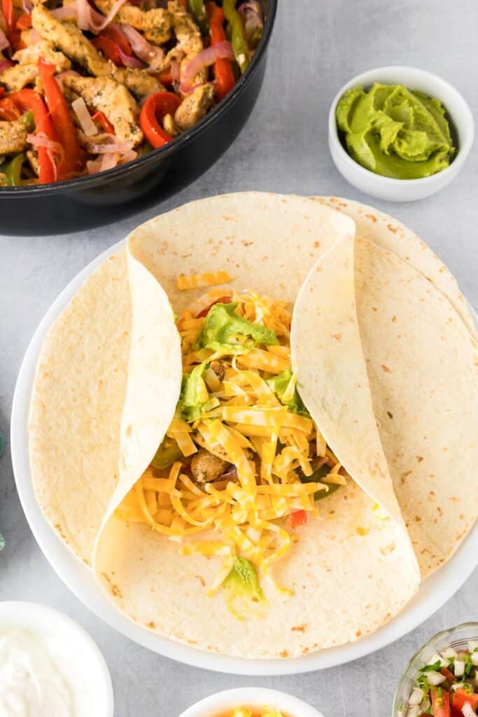 A tortilla filled with chicken fajita filling and cheese being folded on the left and right sides to be rolled.