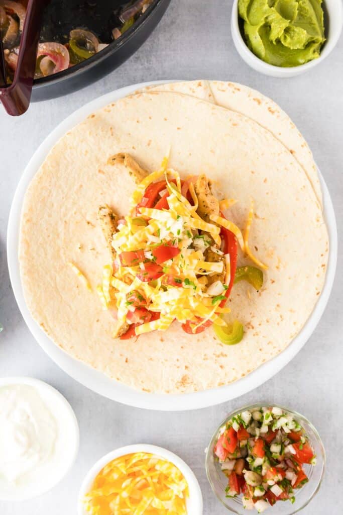 A tortilla on a plate filled with chicken fajita filling, tomatoes and cheese ready to be rolled.