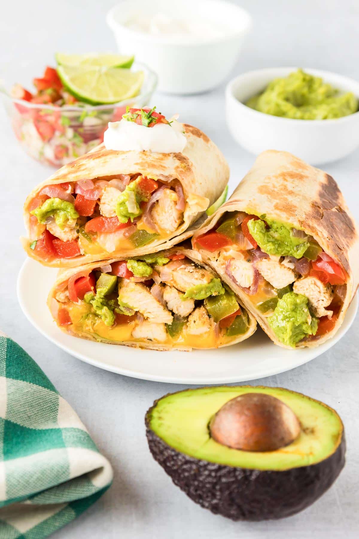 A serving platter with three chicken fajita wraps stacked on the plate cut so you can see the filling with guacamole, avocado, pico de gallo and sour cream on the table nearby.