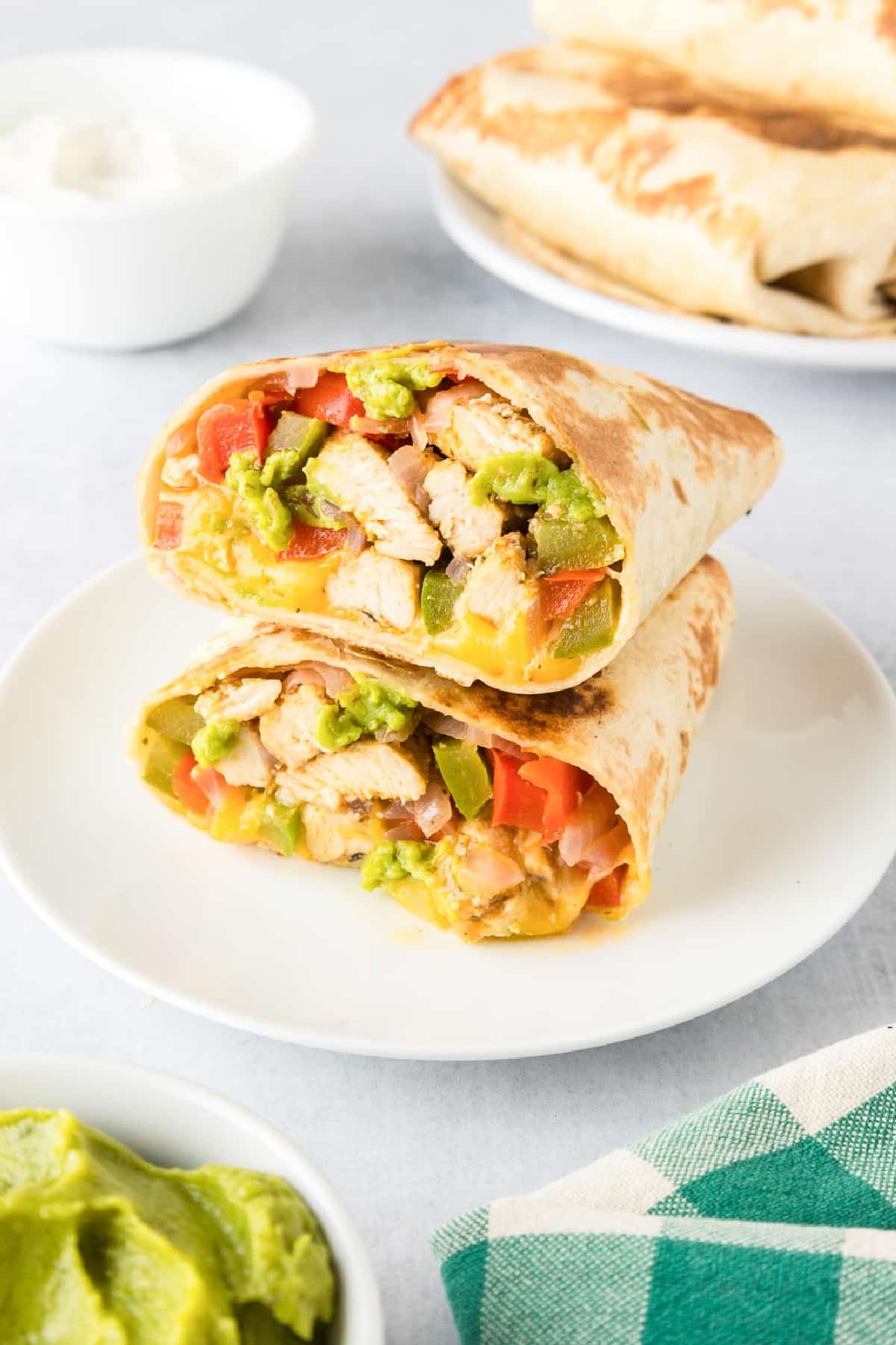 A plate with two burrito halves filled with chicken fajita mixture stacked on a plate so you can see the filling with more wraps and toppings in the background on the table.