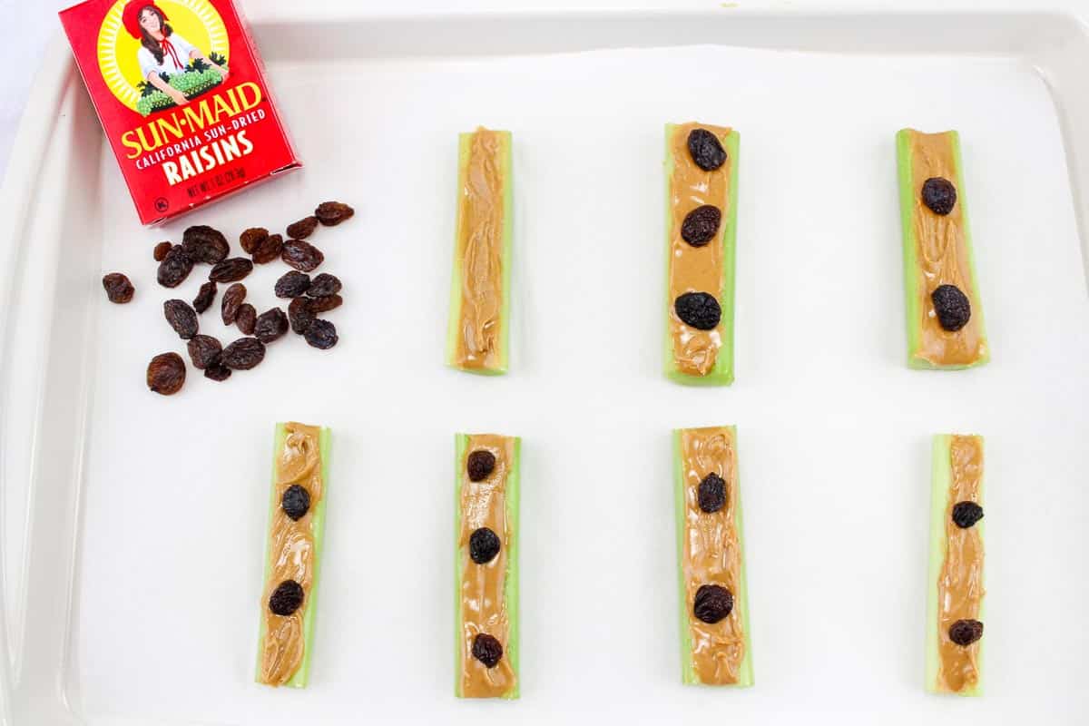 Celery sticks topped with peanut butter and raisins, arranged on a white tray with a box of raisins nearby.