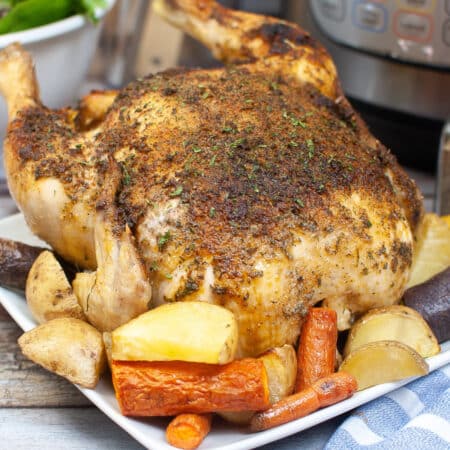 A roasted chicken seasoned with herbs and spices is served on a white plate with carrots and potatoes in front of an instant pot.