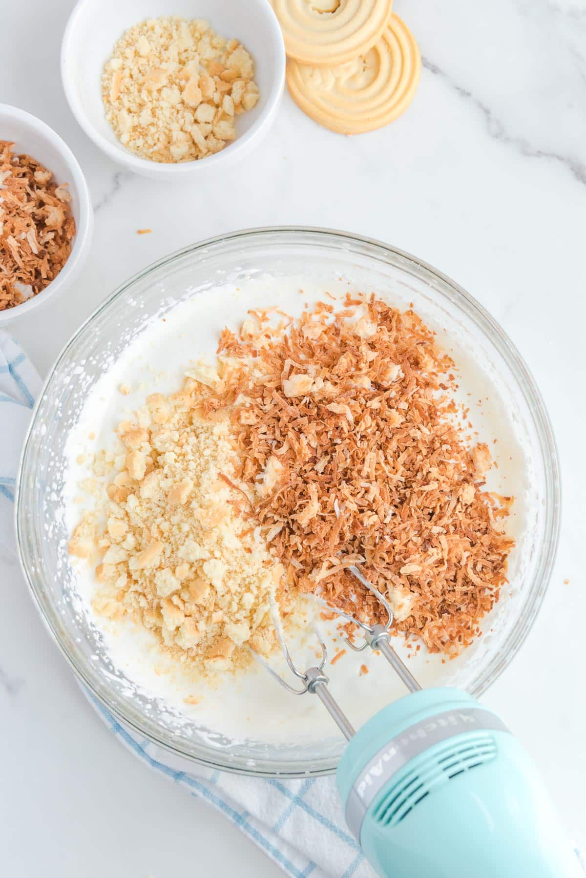 A mixing bowl full of a creamy ice cream mixture with toasted coconut and shortbread cookie crumbs being mixed in.