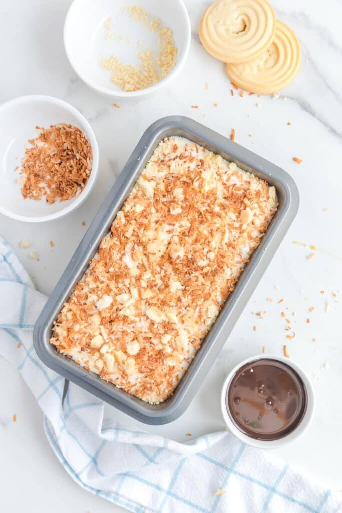 A loaf pan filled with samoa ice cream mixture topped with toasted coconut surrounded by more samoa cookie toppings.