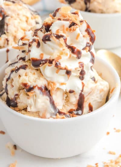 A bowl of samoa ice cream topped with whipped cream, chocolate syrup, and crunchy coconut with a spoon next to the bowl.