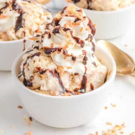 A bowl of samoa ice cream topped with whipped cream, chocolate syrup, and crunchy coconut with a spoon next to the bowl.