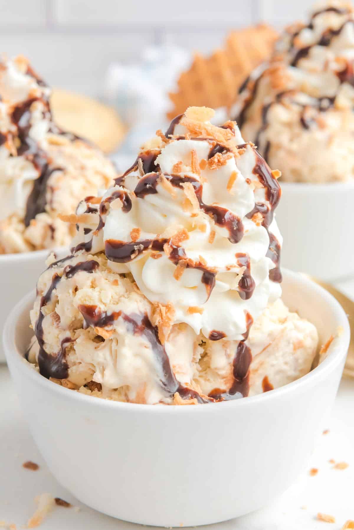 Bowl of samoa ice cream topped with whipped cream, chocolate syrup, and sprinkled coconut flakes with more bowls of ice cream in the background.