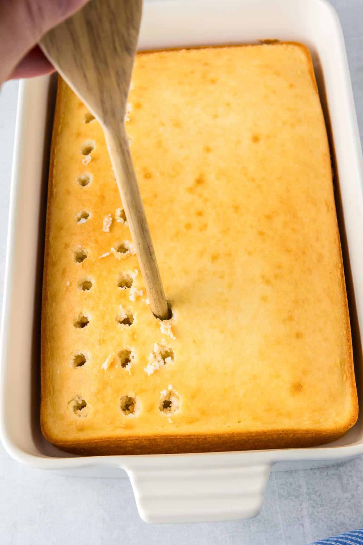 A rectangular cake in a white baking dish being poked with holes with the end of a wooden spoon in rows.