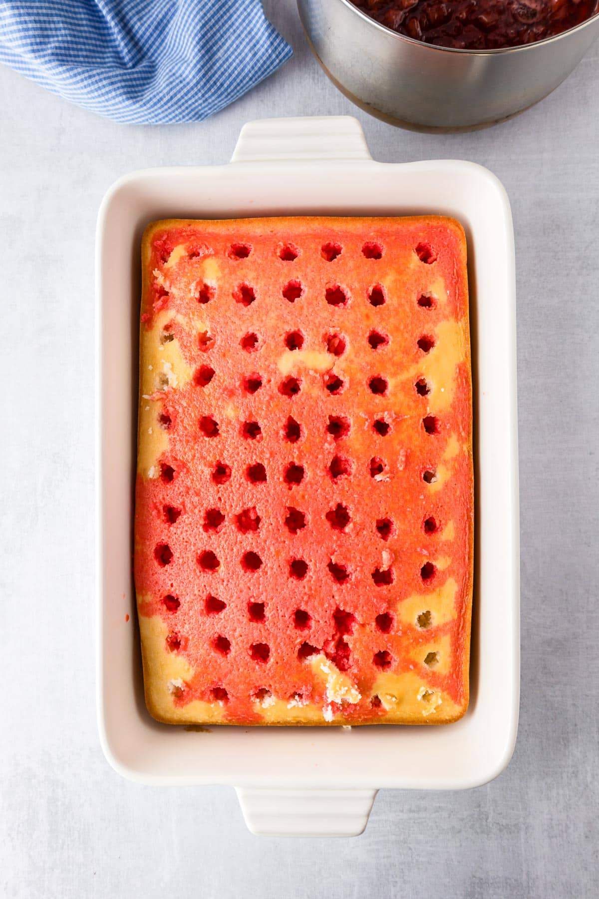 Baking dish with a rectangular cake with evenly space holes in the cake and a red strawberry sauce poured over the cake.
