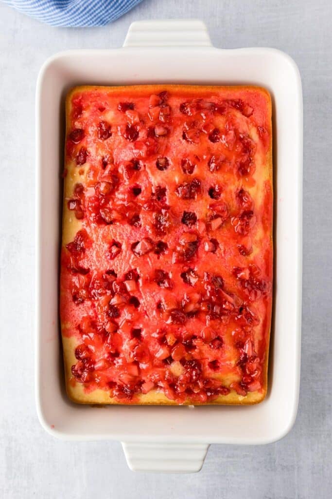 Baking dish with a rectangular cake with evenly space holes in the cake and a real pieces of cooked strawberries over the cake.