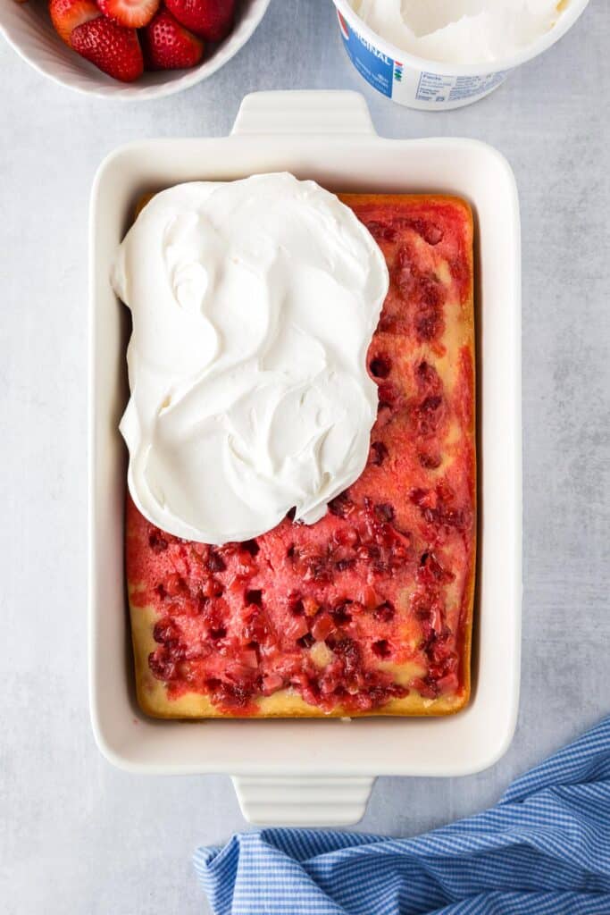 A rectangular dessert in a baking dish with strawberry poke cake full of holes and strawberry sauce, with a whipped topping being spread on top of the cake from overhead.