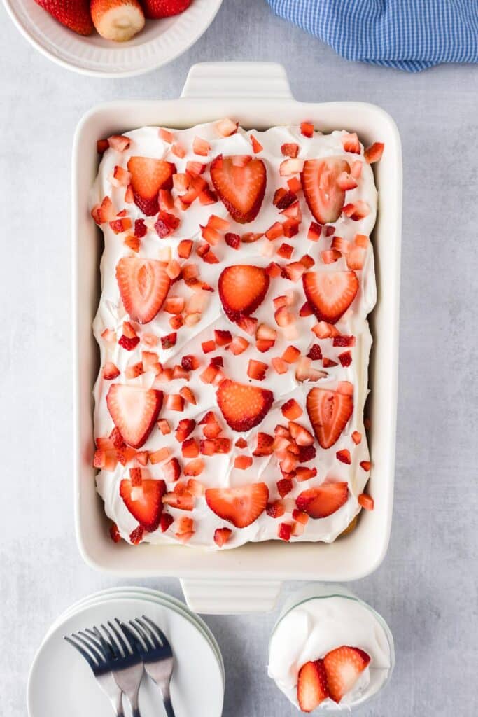 A rectangular dish filled with strawberry poke cake, frosting and strawberry pieces on a table with plates and forks to serve.