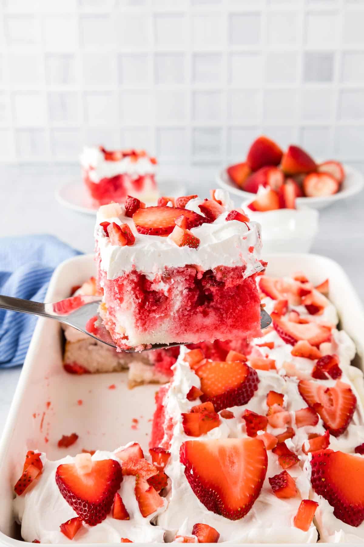 A piece of red and white strawberry poke cake topped with whipped cream and strawberry slices being lifted from a baking dish.