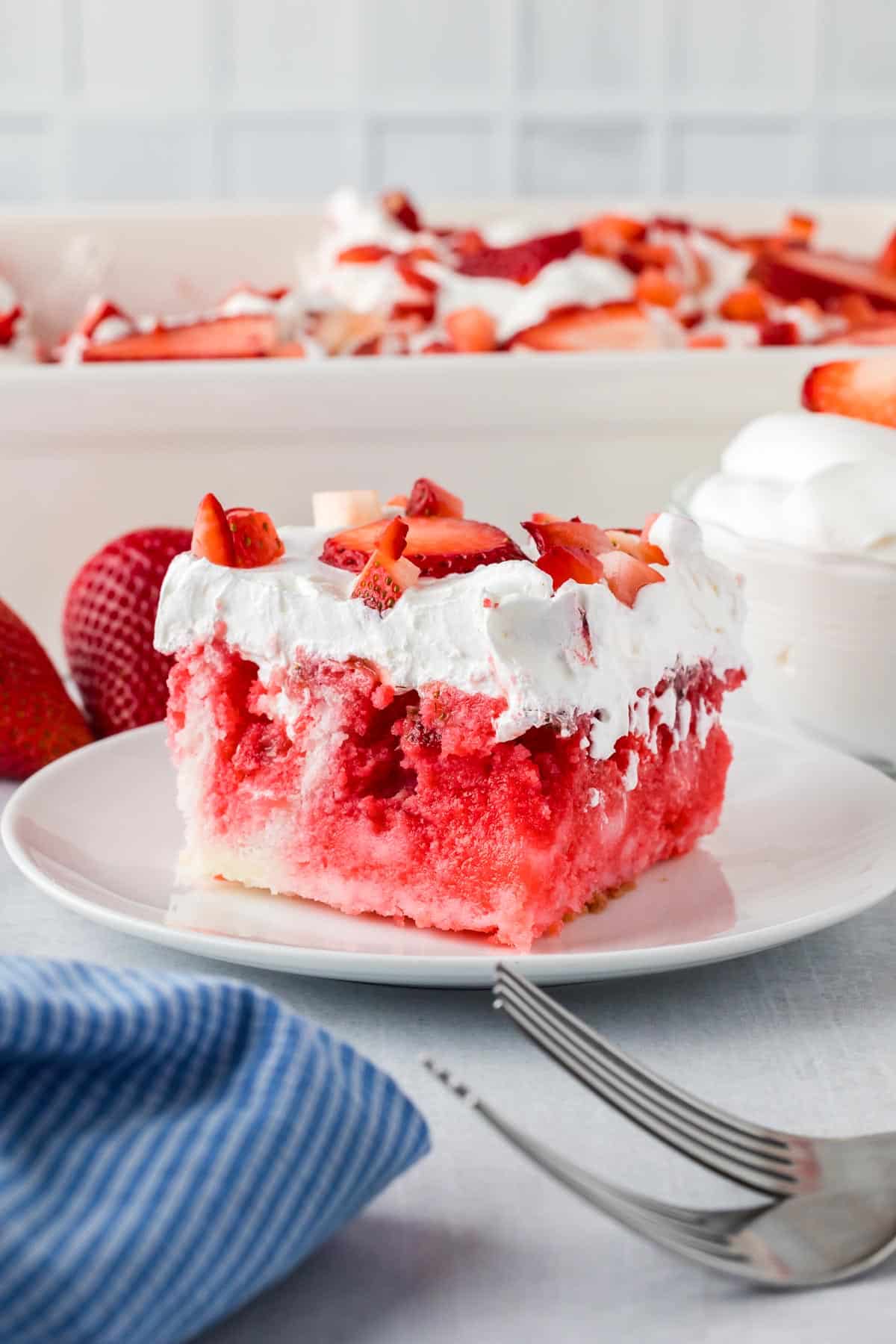 A slice of strawberry JELLO poke cake topped with whipped cream and diced strawberries on a white plate with more cake in a pan in the background on a kitchen table.