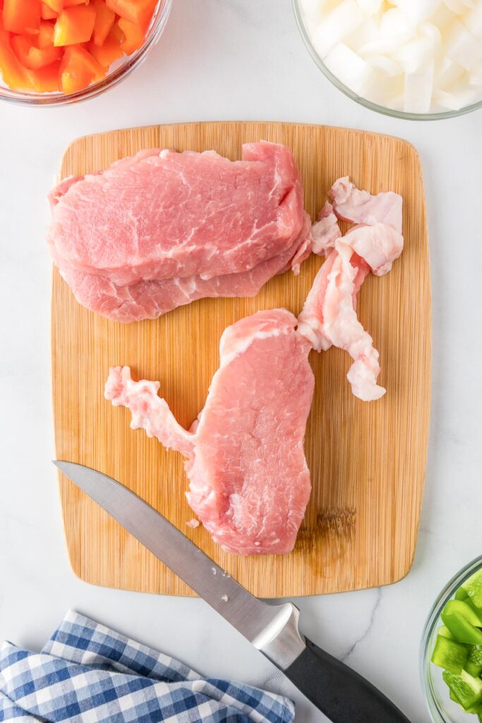 Two raw pork chops placed on a cutting board with the fat being trimmed with a knife.