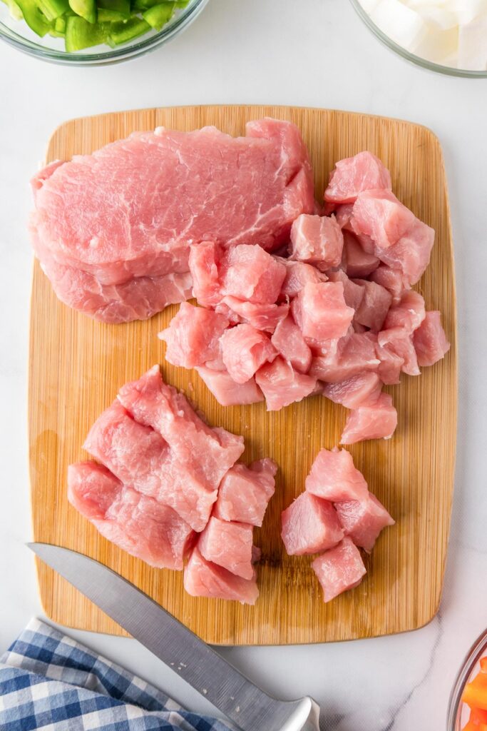 Raw boneless pork chops being sliced on a cutting board into cubes with a knife.