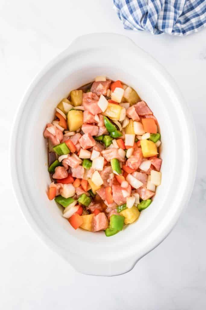 A white oval dish slow cooker filled with raw boneless pork chop pieces, chopped peppers, onions and pineapple mixed together.