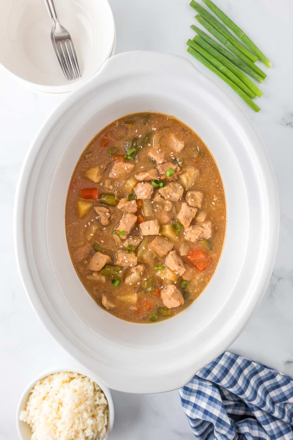 A slow cooker filled with sweet and sour pork and vegetables with a bowl of rice and a bowl with a fork nearby.