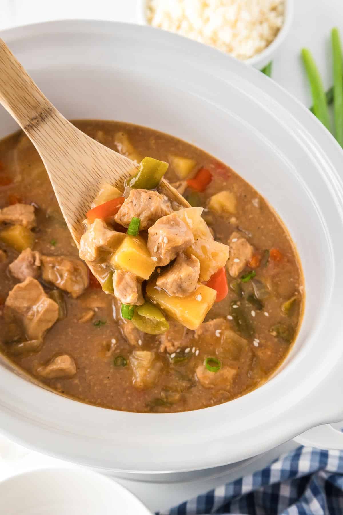 A spoonful of sweet and sour pork mixed with pineapple and peppers being lifted from a slow cooker.