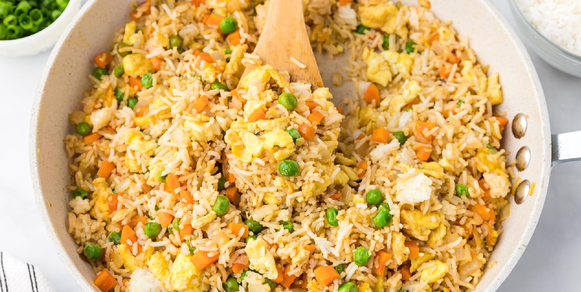 A pan filled with fried rice with a wooden spoon scooping it full of egg, peas, carrots and other vegetables.