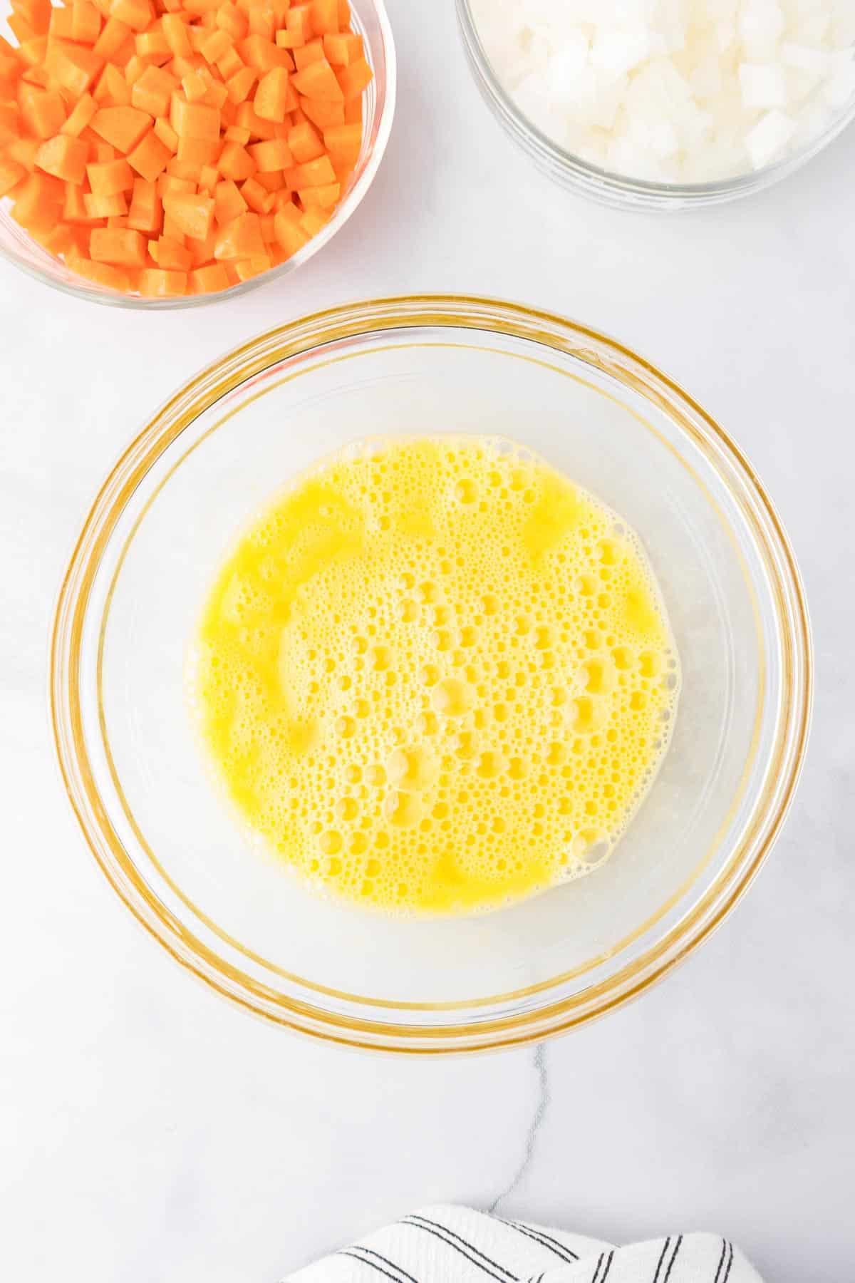 A bowl of beaten eggs with diced carrots and onions in bowls nearby.