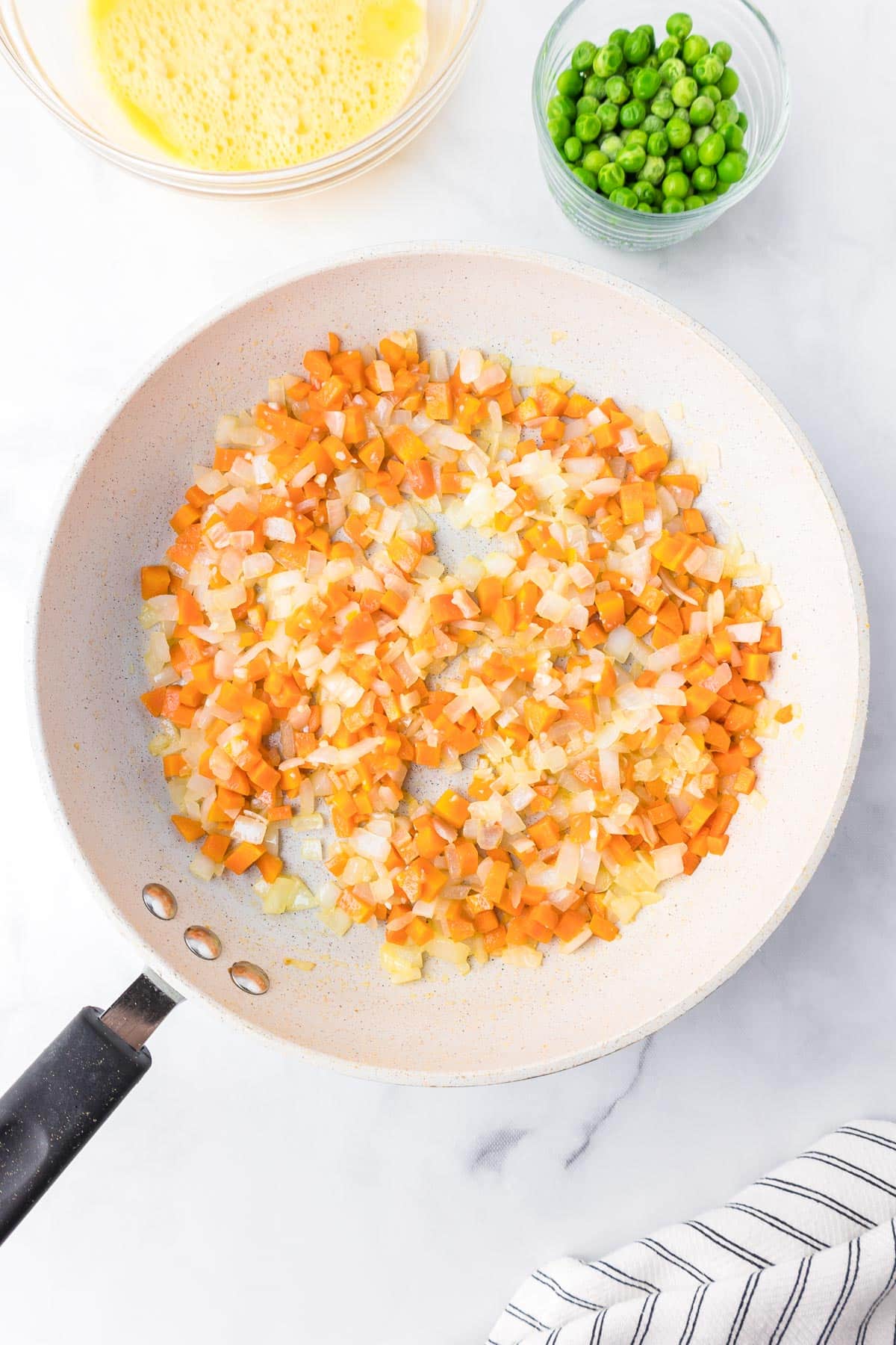 A frying pan containing sautéed diced carrots and onions with peas and beaten liquid eggs in bowls nearby.