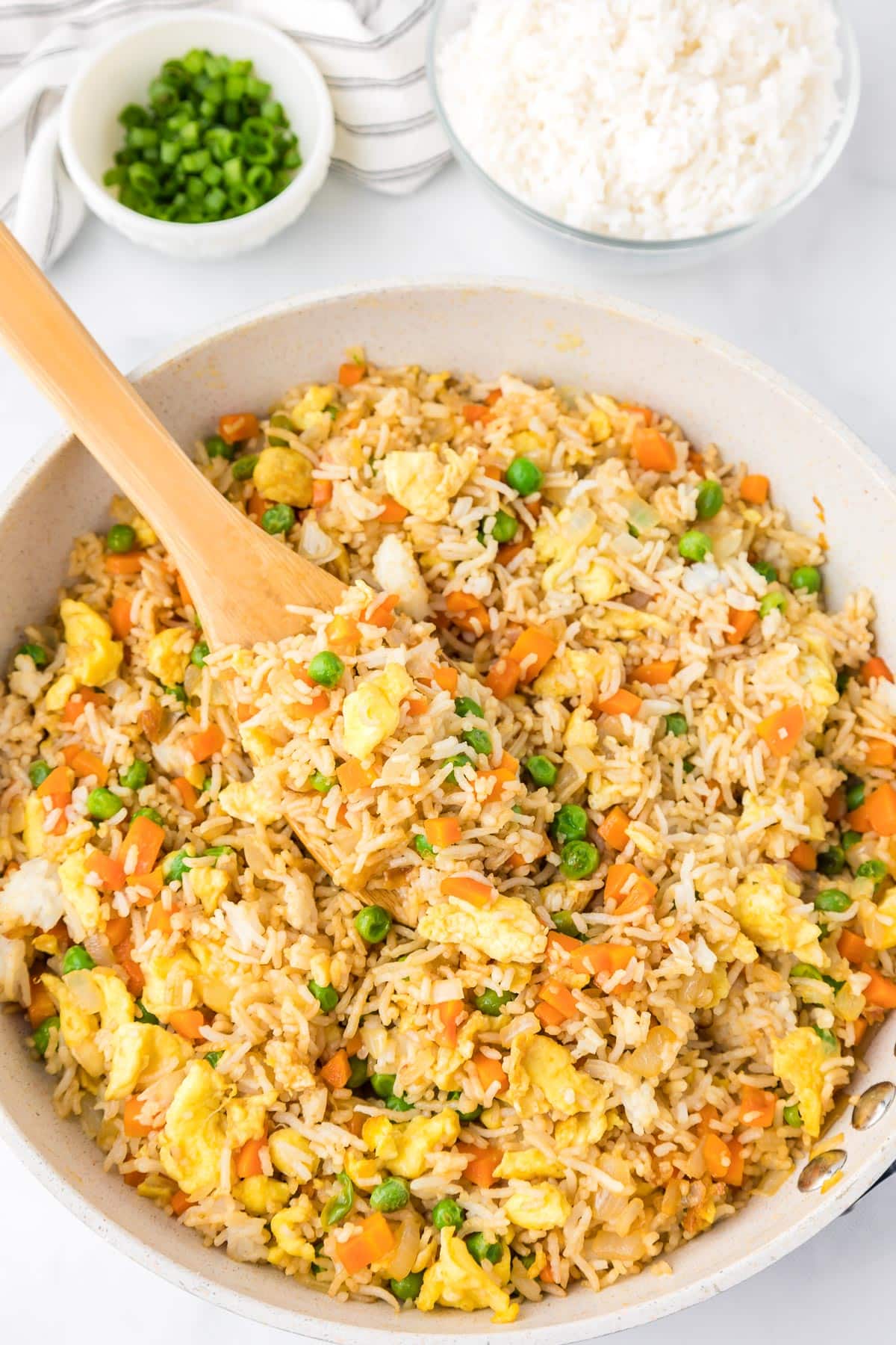 A skillet filled with fried rice containing scrambled eggs, peas, carrots, and green onions being scooped by a wooden spoon.