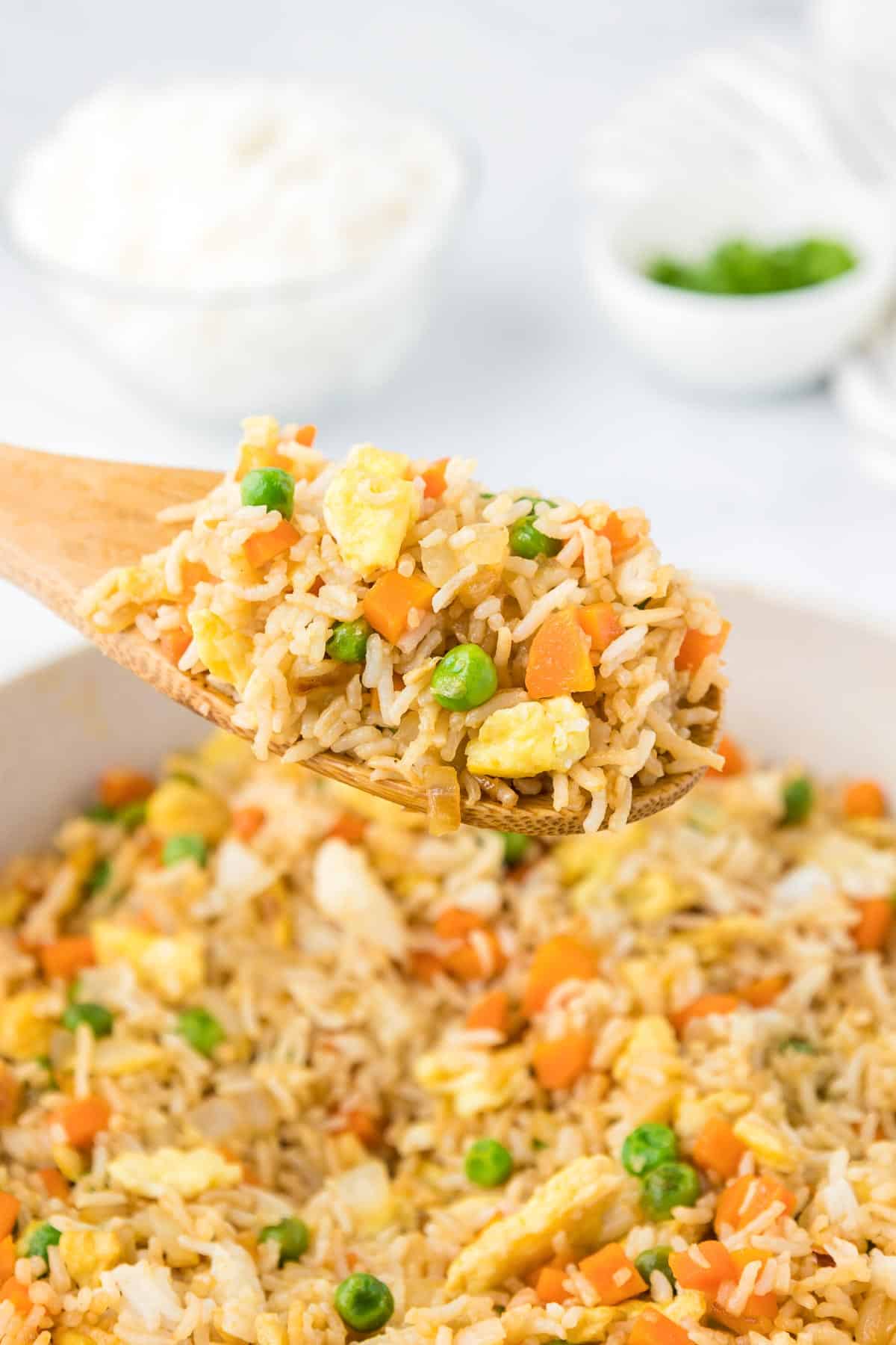 A wooden spoon holding a scoop of fried rice with peas, carrots, and scrambled egg above a pan filled with fried rice.