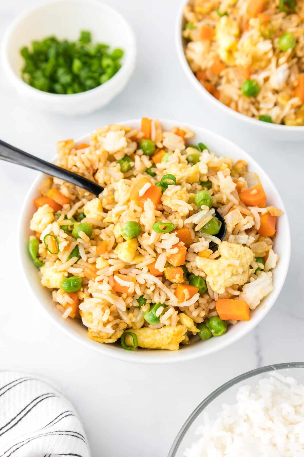 A bowl fill of fried rice with peas, carrots, and scrambled eggs with a spoon and more fried rice and chopped green onions on the table nearby.