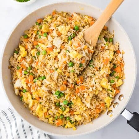 A skillet filled with fried rice mixed with peas, carrots, and scrambled eggs with a wooden spoon stirring.