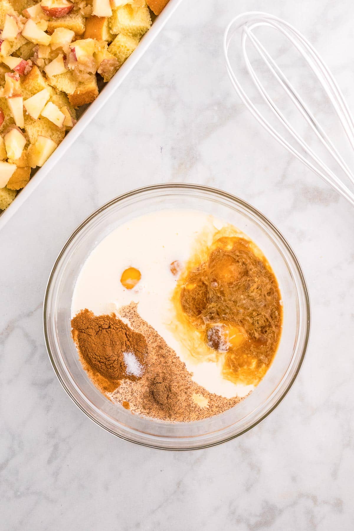 A glass bowl containing milk, eggs, and various spices with a whisk nearby on the counter to mix for apple french toast casserole.