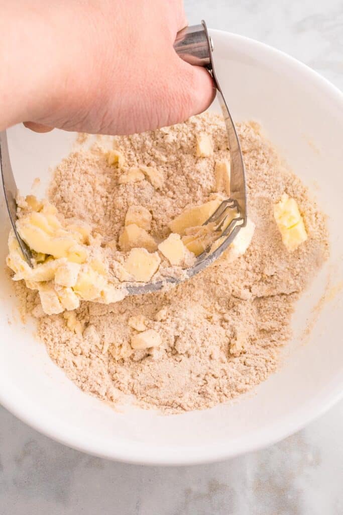 A hand using a pastry cutter to mix flour and butter in a white bowl for the apple french toast casserole topping.