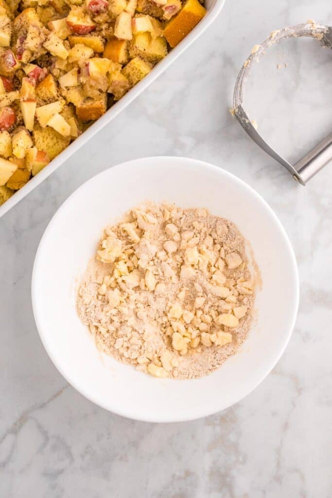 A white bowl contains a crumbly mixture of flour and butter being mixed together with a metall pastry tool on the counter and the apple french toast casserole nearby.