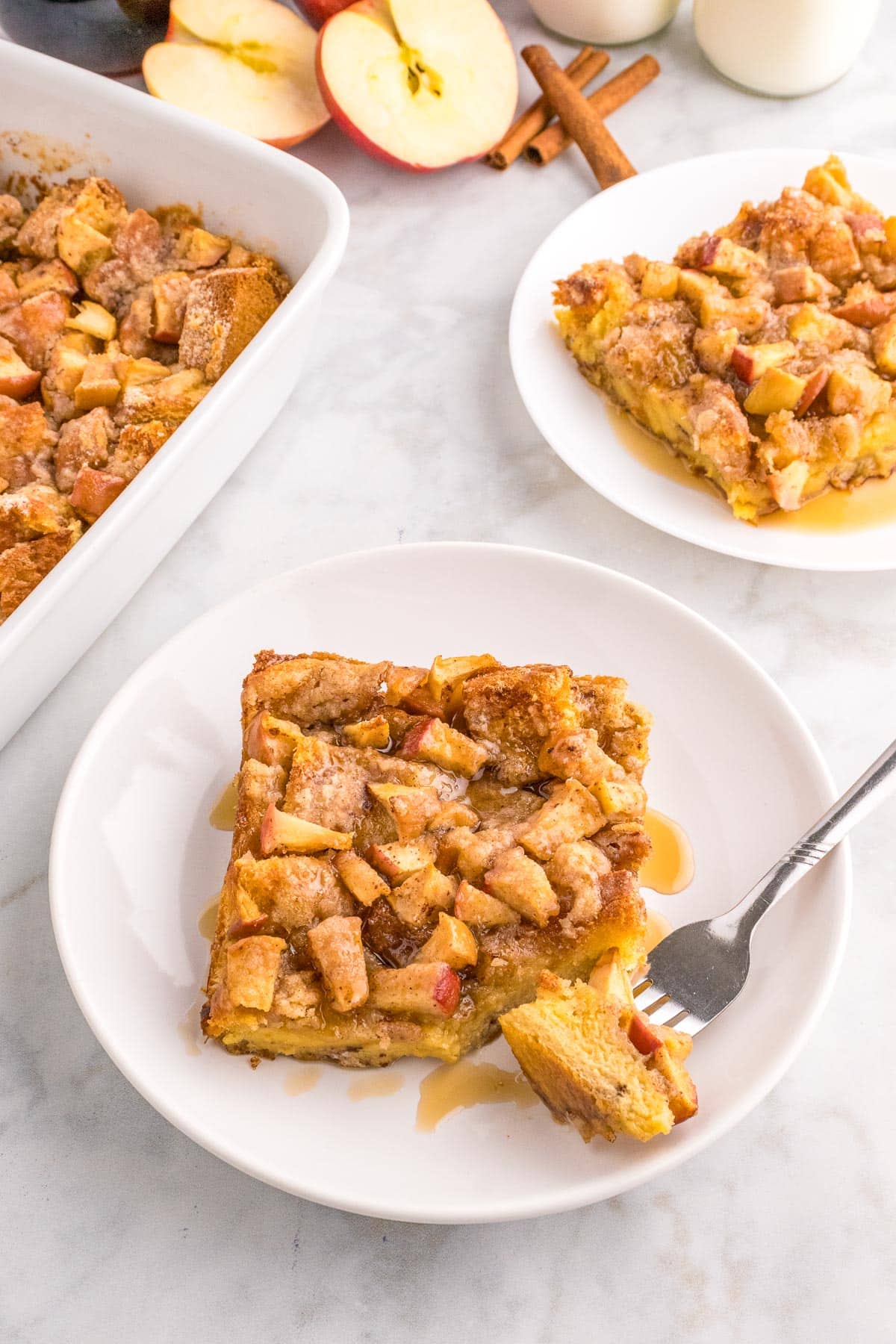 A square slice of apple French toast casserole on a plate from overhead with a fork taking a bite, with a second plate and the full casserole dish in the background on the table.