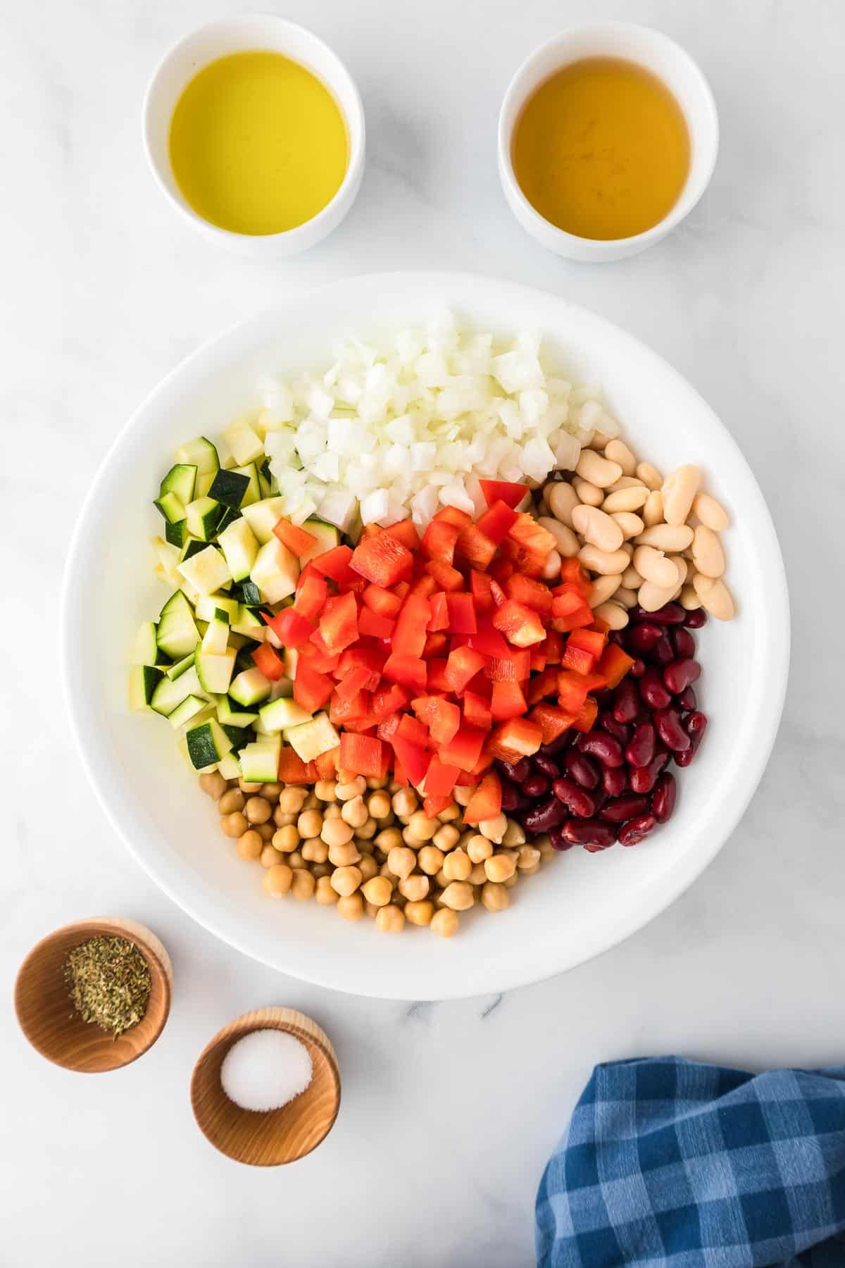 A bowl with chopped vegetables for bean chickpea salad and ingredients for the dressing in bowls nearby.