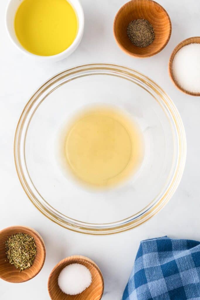 A large glass bowl filled with apple cider vinegar with other herbs, spices and oil in small bowls nearby on the counter.