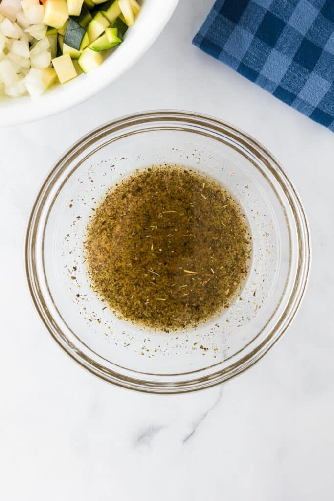 A glass bowl filled with and herb filled dressing for bean chickpea salad with the bowl of chopped vegetables nearby.