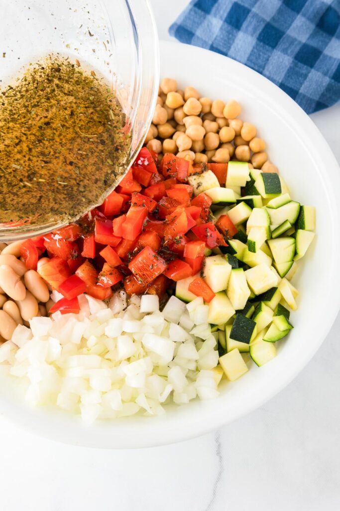 A bowl of all the vegetables for chickpea bean salad with a herb filled dressing being poured over the salad.