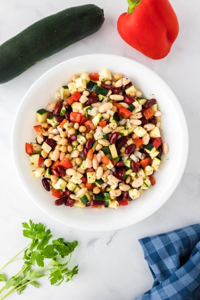 A white bowl filled with a finished chickpea bean salad with more ingredients on the counter nearby.